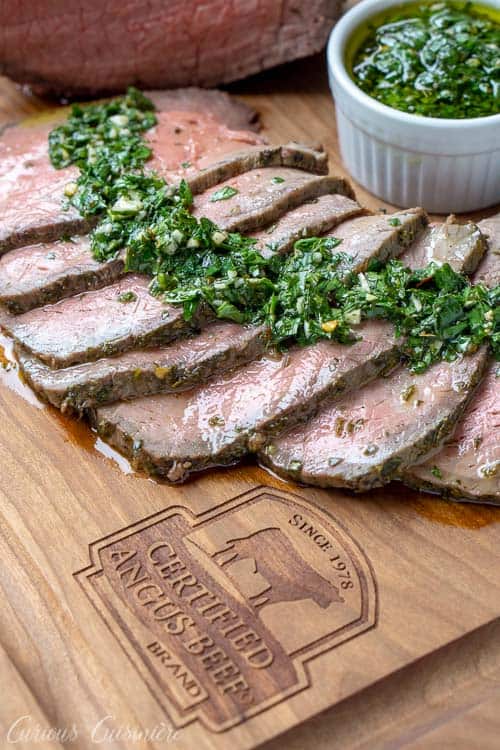 Sliced top round roast with chimichurri sauce on a Certified Angus Beef Brand branded wood cutting board. 