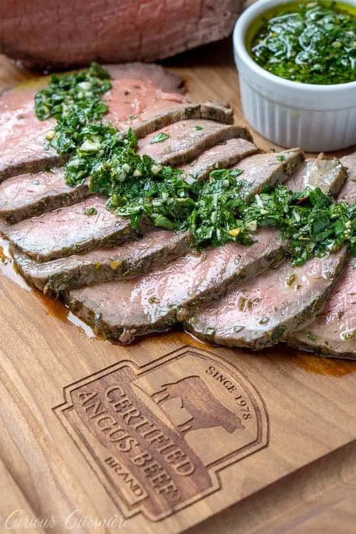 Sliced top round roast with chimichurri sauce on a Certified Angus Beef Brand branded wood cutting board.