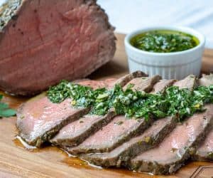 Horizontal shot of a chimichurri marinated top round beef roast. Served with a side of chimichurri.