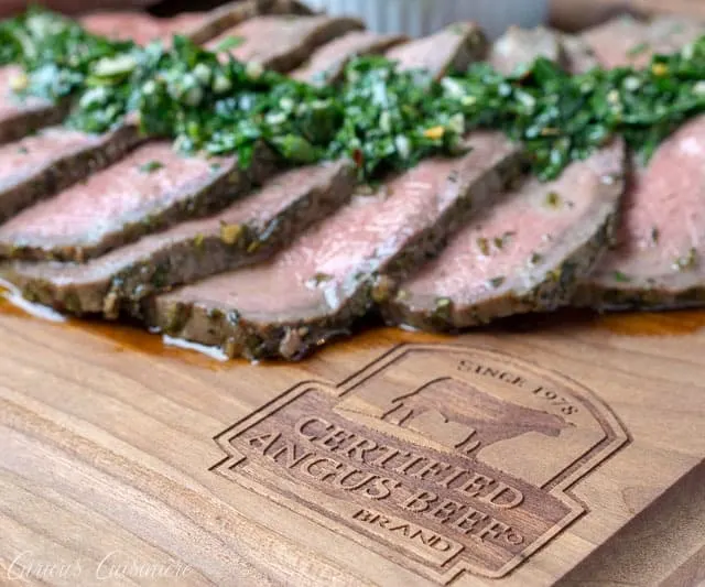 Sliced top round roast with chimichurri sauce on a Certified Angus Beef Brand branded wood cutting board.