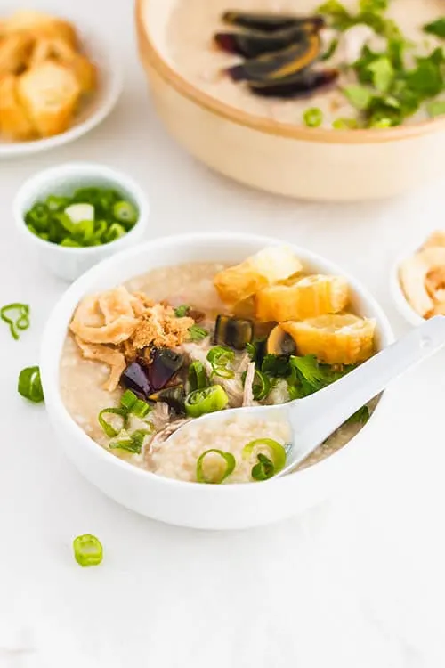 A spoon in a bowl of century egg and pork congee with green onions and fried yu tiao Chinese donuts. | www.CuriousCuisiniere.com 