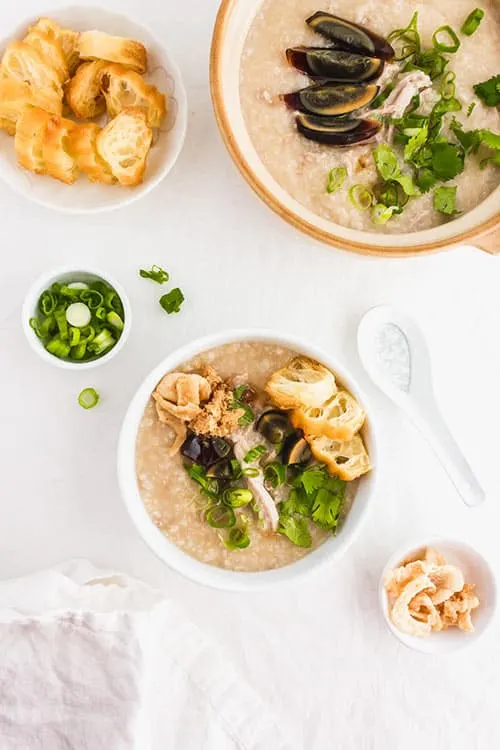 A pot and a bowl of preserved century egg congee with green onions and fried yu tiao Chinese donuts and green onions. | www.CuriousCuisiniere.com