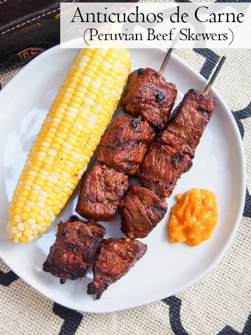 Overhead image of Anticuchos, Peruvian grilled beef skewers on a plate with corn and sauce
