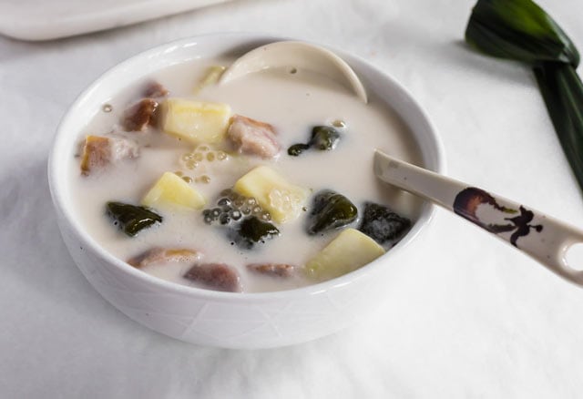 Bubur Cha Cha, a creamy and sweet Malaysian dessert made from yam, taro, and coconut milk in a white bowl with a decorative spoon and pandan leaf. 