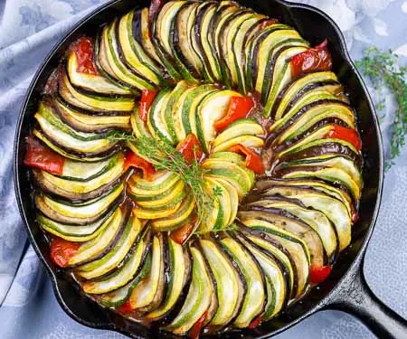 Overhead image of layered ratatouille in a cast iron skillet with eggplant, summer squash, zucchini, tomatoes, and red peppers.