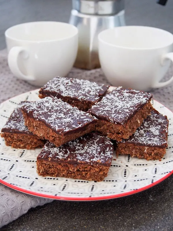 Stacked chocolate slice topped with flaked coconut with tea mugs